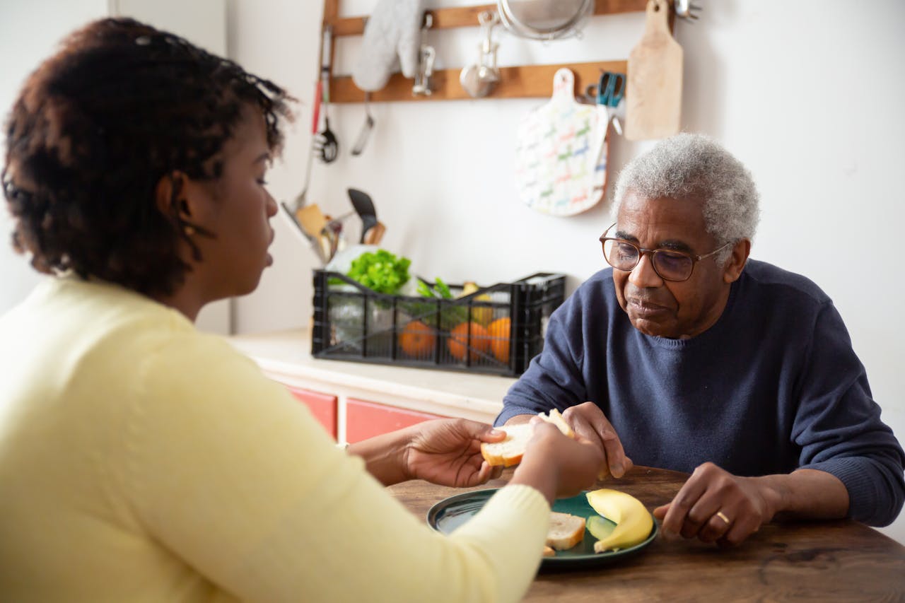 A woman assists an elderly man with meals at home, showcasing care and support.